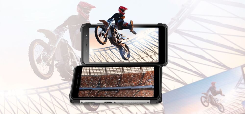Obraz przedstawia smartfon z wyświetlaczem, na którym widoczny jest motocyklista w powietrzu. W tle znajduje się rozmyty obraz motocyklisty na stadionie.