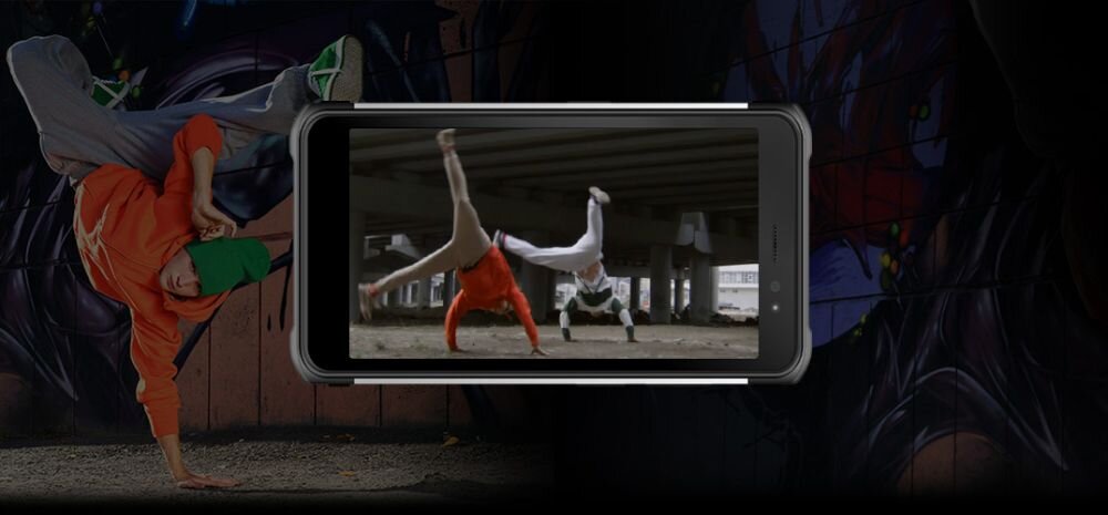 Na obrazie widoczny jest smartfon z wyświetlaczem, na którym pokazane są dwie osoby wykonujące akrobacje pod mostem. W tle znajduje się graffiti na ścianie oraz osoba w pomarańczowej bluzie i zielonej czapce wykonująca taniec uliczny.