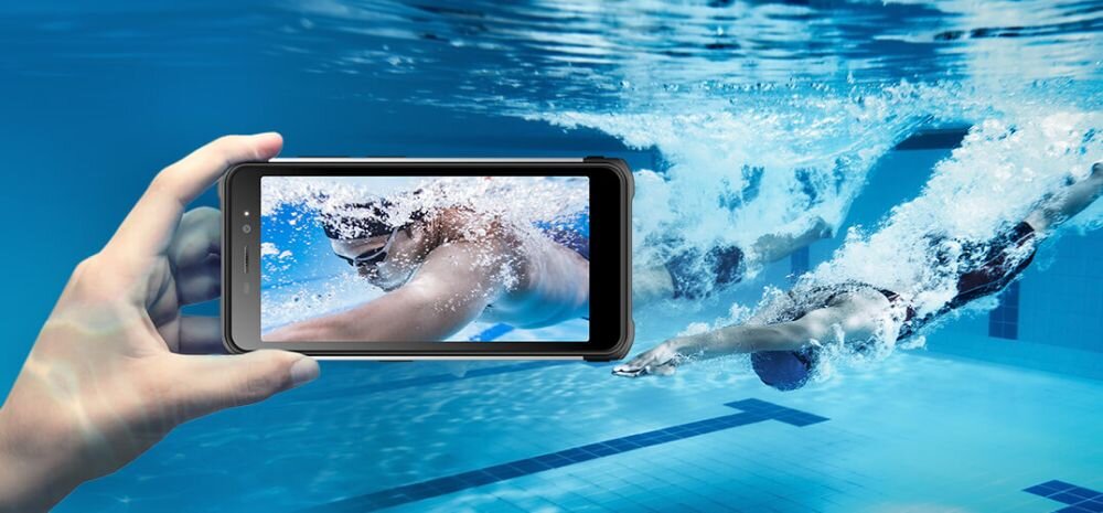 Osoba trzyma smartfon, na ekranie którego widać pływaka pod wodą. W tle znajduje się basen z pływającymi osobami.