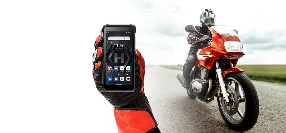 Osoba w rękawicach motocyklowych trzyma smartfon z widocznym ekranem, na którym jest godzina 11:56 i kilka ikon aplikacji. W tle jedzie motocyklista na czerwonym motocyklu.