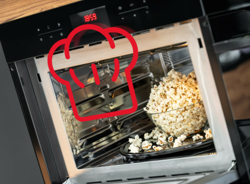 Otwarta kuchenka mikrofalowa z wyświetlaczem pokazującym godzinę 18:59, wewnątrz której znajduje się miska z popcornem. Na drzwiach kuchenki widoczny jest czerwony symbol czapki kucharskiej.