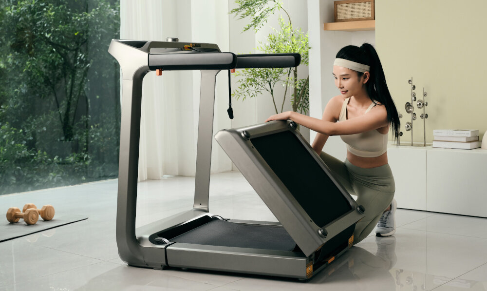 Kobieta w sportowym stroju składa bieżnię elektryczną w jasnym, nowoczesnym wnętrzu. W tle widoczne są rośliny i hantle na podłodze.