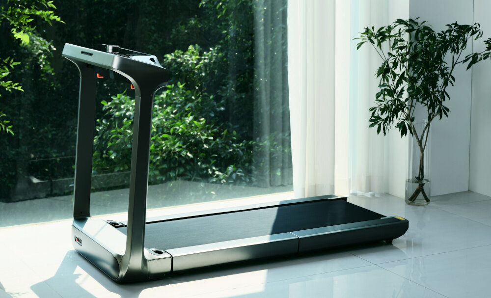 Na obrazie widoczna jest bieżnia elektryczna ustawiona w jasnym pomieszczeniu z dużymi oknami i rośliną w doniczce. Bieżnia ma nowoczesny design i ciemną kolorystykę.