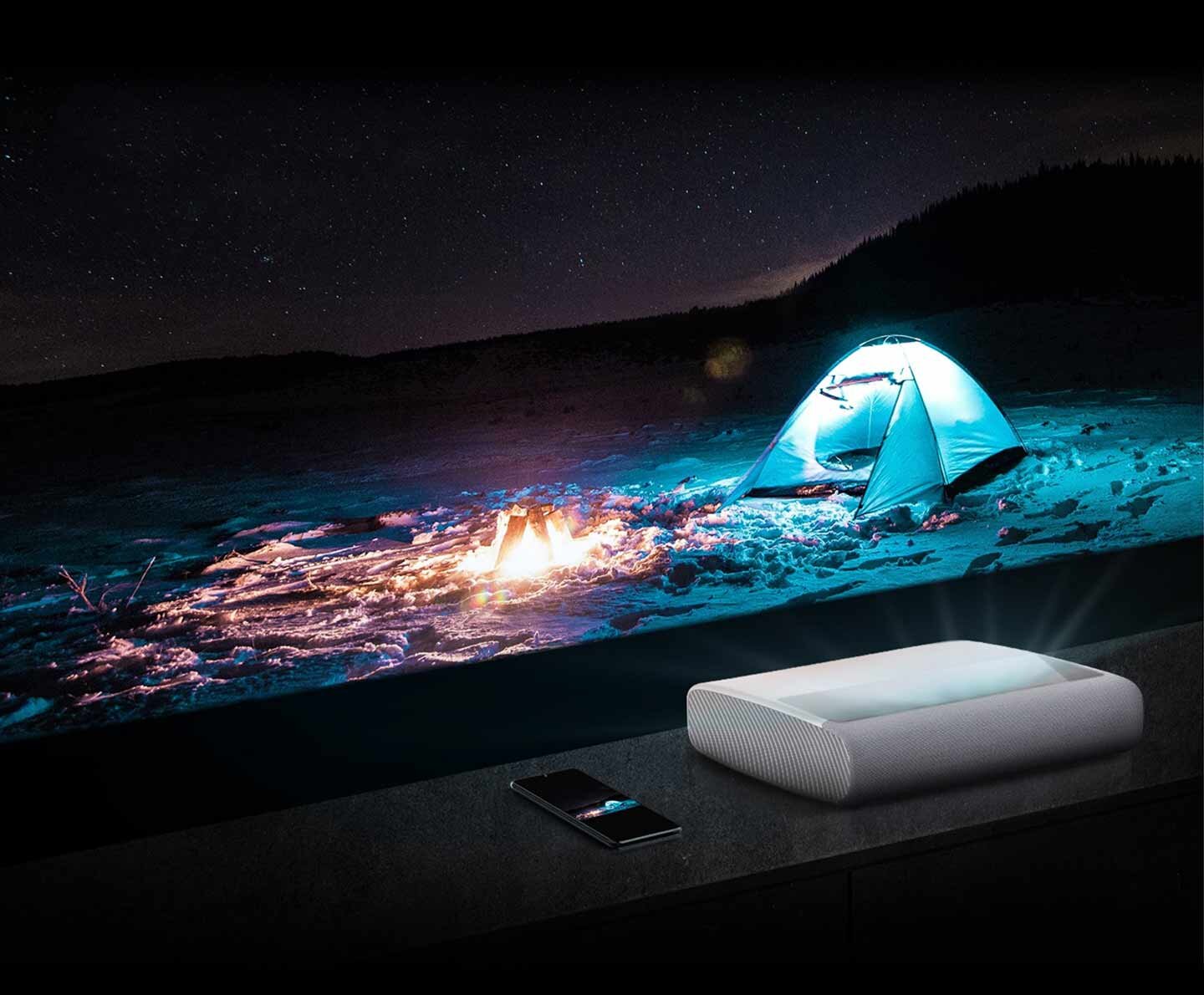 Na obrazie widać projektor wyświetlający scenę z namiotem i ogniskiem na tle nocnego nieba. Obok projektora leży smartfon.