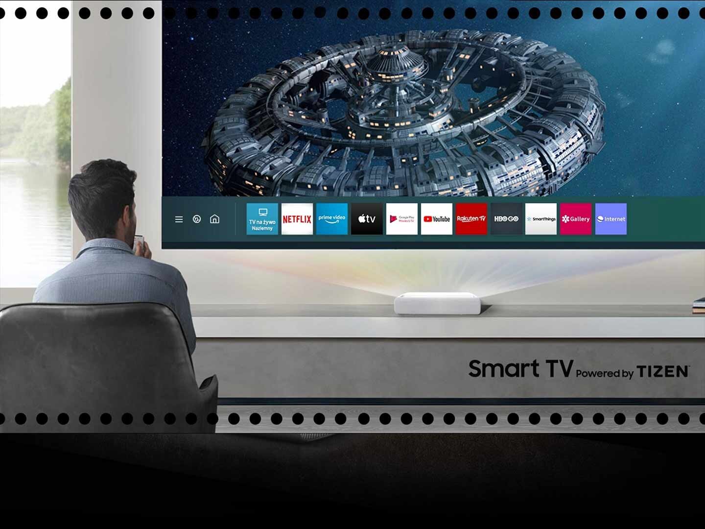 Osoba siedzi przed dużym ekranem, na którym wyświetlany jest obraz kosmicznego statku oraz ikony aplikacji takich jak Netflix, Prime Video, YouTube. Na dolnej części ekranu widnieje napis \'Smart TV Powered by TIZEN\'.