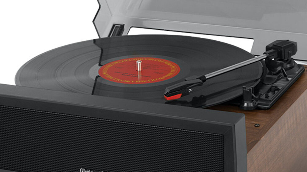 Gramofon z drewnianą obudową odtwarza czarną płytę winylową, z widoczną igłą na ramieniu. Pokrywa gramofonu jest otwarta, a na płycie znajduje się czerwono-żółta etykieta.