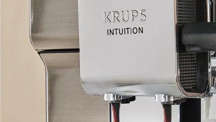 Zbliżenie na ekspres do kawy z widocznym napisem \'KRUPS INTUITION\'. Z urządzenia wypływa kawa.
