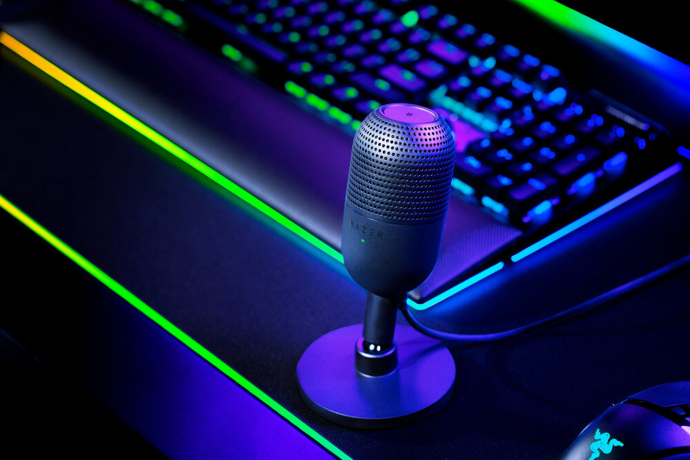 Na obrazie znajduje się mikrofon na biurku z podświetlaną klawiaturą w tle. Na mikrofonie widoczne jest logo \'RAZER\'.