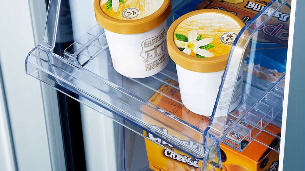 Wnętrze lodówki z przezroczystą półką, na której znajdują się dwa pojemniki z lodami o złotych pokrywkach i opakowanie z napisem \'Cheese\'.