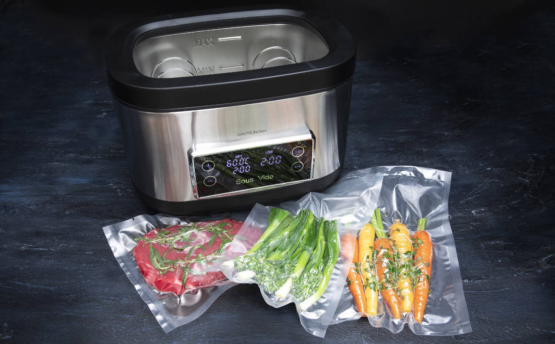 Urządzenie do gotowania Sous Vide marki GASTRONOMA z wyświetlaczem pokazującym temperaturę i czas. Przed urządzeniem znajdują się trzy zapakowane próżniowo porcje jedzenia: mięso, zielone warzywa i marchewki.