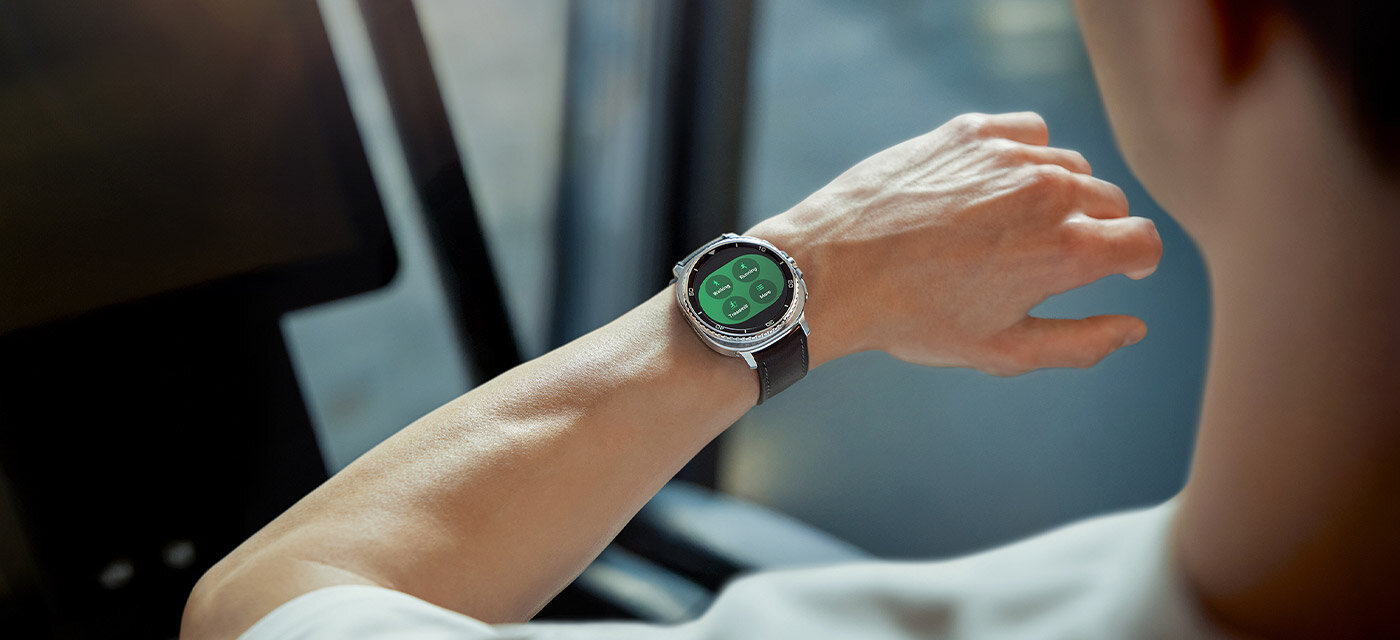 Osoba nosząca Galaxy Watch8 Classic. Funkcja śledzenia ćwiczeń jest wyświetlana na ekranie, wyświetlając chodzenie, bieganie, jazdę na rowerze i więcej. Osoba patrzy na smartwatch na nadgarstku, który ma okrągły wyświetlacz z zielonym tłem i ikonami. W tle widać rozmyty fragment wnętrza pojazdu lub pomieszczenia.