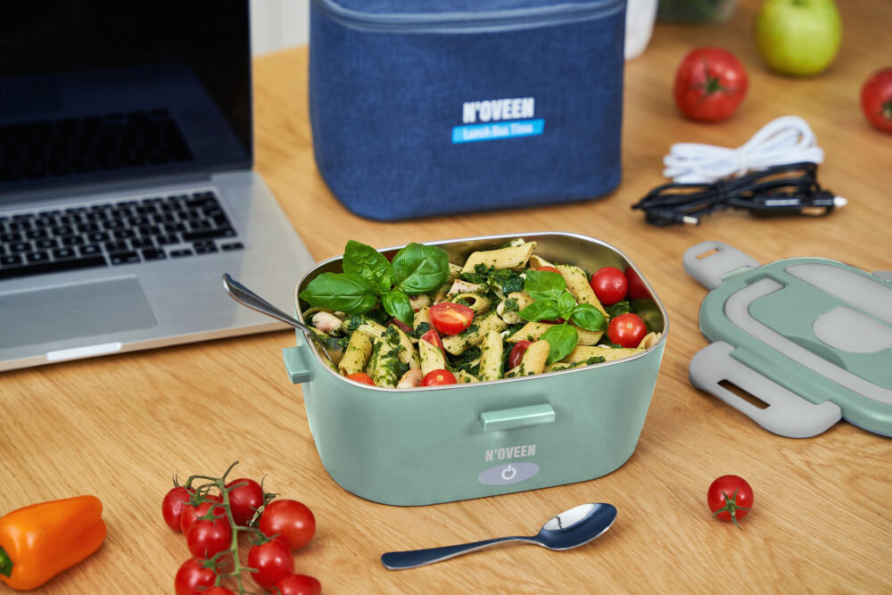 Na drewnianym stole znajduje się elektryczny lunch box NOVEEN wypełniony jedzeniem, obok leży pokrywka i łyżka. W tle widoczna jest torba z napisem \'N\'OVEEN Lunch Box Time\', laptop oraz kilka warzyw.