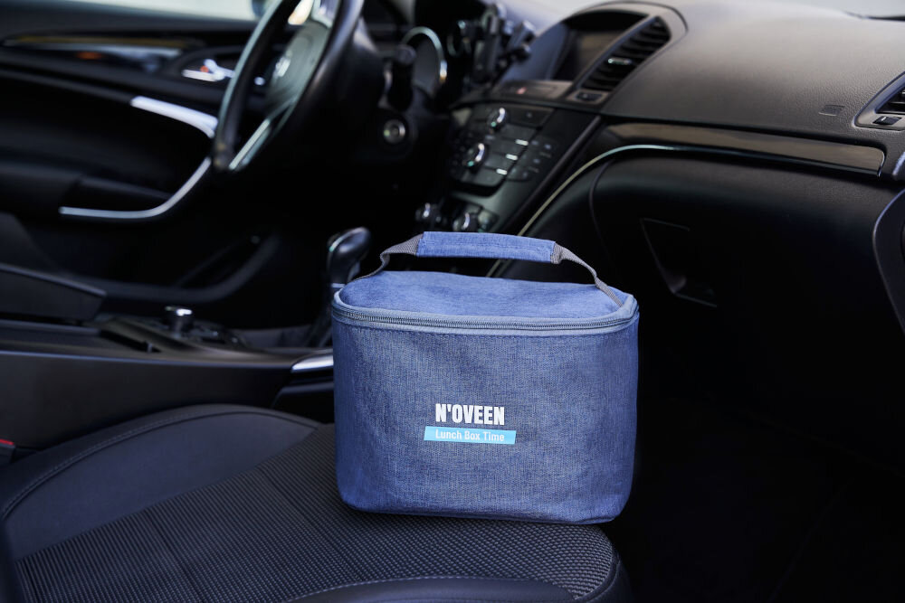 Na siedzeniu samochodu znajduje się niebieska torba z napisem \'N\'OVEEN Lunch Box & More\'. W tle widoczna jest deska rozdzielcza pojazdu.