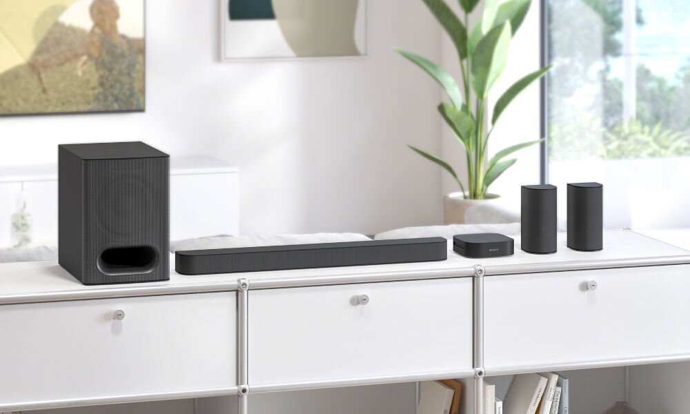 Na białej szafce znajduje się zestaw audio składający się z soundbara, subwoofera i dwóch głośników satelitarnych. W tle widoczna jest roślina doniczkowa i fragment okna.