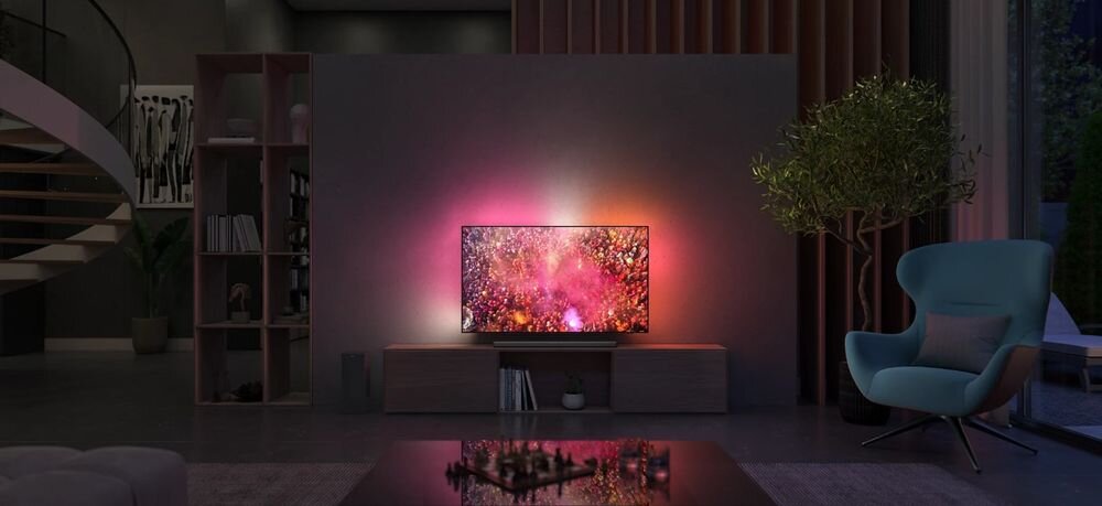 W nowoczesnym salonie znajduje się telewizor wyświetlający kolorowy obraz z efektem świetlnym na ścianie. Obok telewizora stoi szafka z książkami, a w rogu pomieszczenia znajduje się fotel i roślina.
