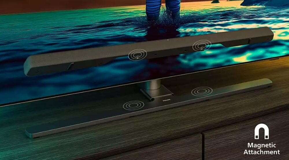 Na obrazie widać szary soundbar umieszczony pod telewizorem, z oznaczeniami miejsc magnetycznego mocowania. W prawym dolnym rogu znajduje się napis \'Magnetic Attachment\' oraz symbol magnesu.