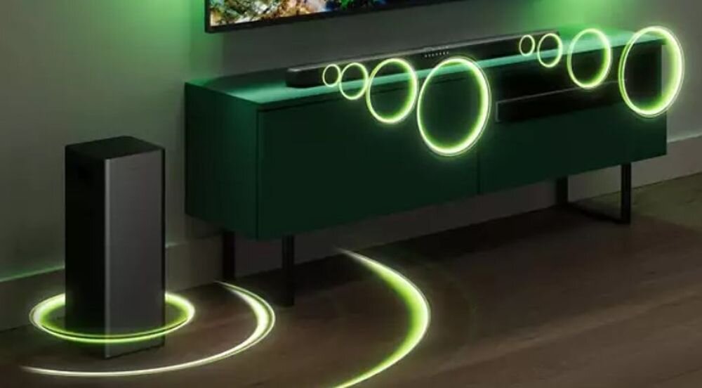 Na obrazie widać szary soundbar umieszczony na ciemnej szafce oraz stojący obok subwoofer. Wokół urządzeń widoczne są zielone, świetliste kręgi sugerujące efekt dźwięku przestrzennego.