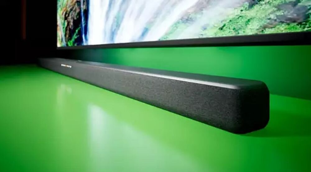 Na zielonym tle znajduje się szary soundbar umieszczony pod telewizorem. Na soundbarze widoczne jest logo producenta.