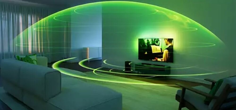 W salonie znajduje się telewizor z włączonym obrazem oraz soundbar, a wokół nich widoczna jest zielona, świetlna wizualizacja dźwięku przestrzennego. Pomieszczenie jest nowoczesne, z sofą i fotelem.