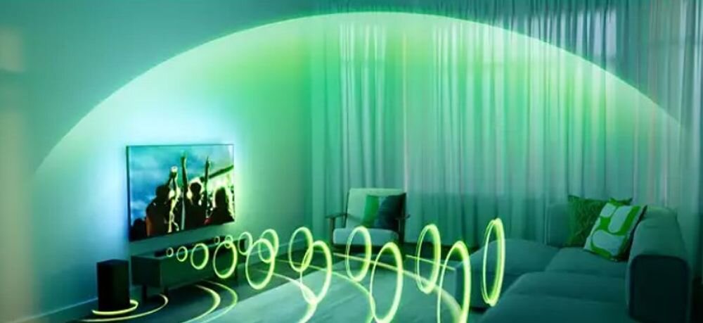 Pokój z telewizorem na ścianie, przed którym znajduje się soundbar i subwoofer. Zielone fale dźwiękowe wizualizują efekt dźwięku przestrzennego.
