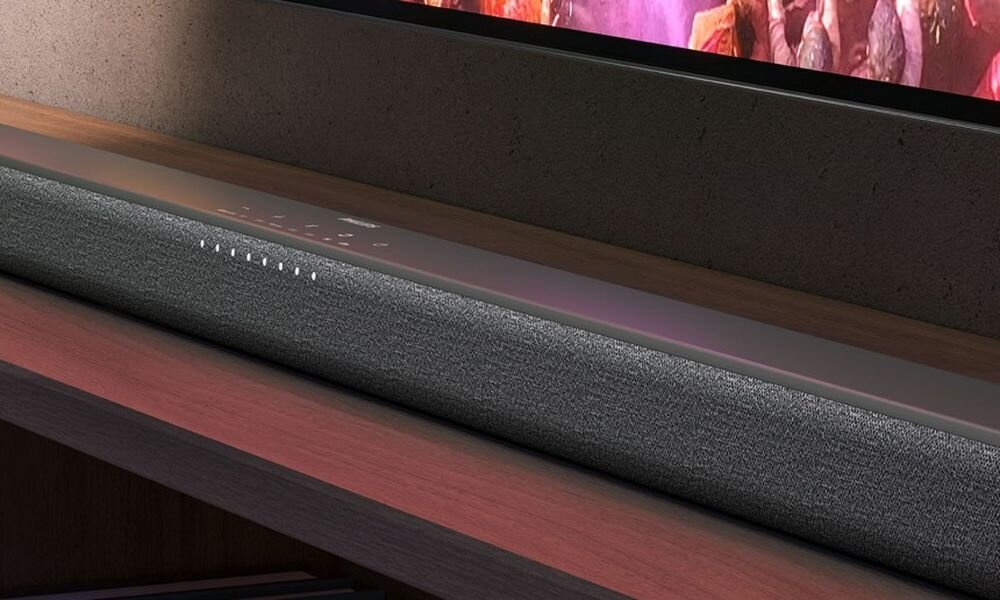 Na obrazie widoczny jest szary soundbar umieszczony na drewnianej półce, z wyświetlaczem LED pokazującym różne ikony i wskaźniki. W tle znajduje się fragment telewizora z kolorowym obrazem.