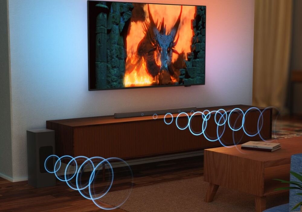 Na obrazie widać telewizor z wyświetlonym smokiem w ogniu oraz soundbar umieszczony na szafce pod telewizorem. Obok znajduje się subwoofer, a wokół soundbara i subwoofera widoczne są graficzne fale dźwiękowe.