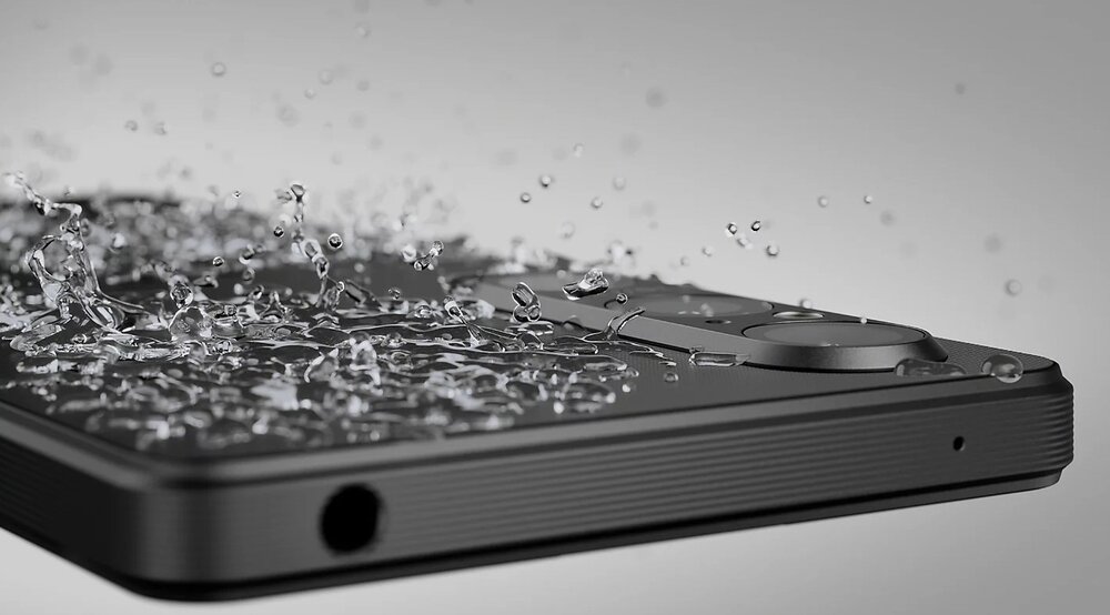Smartfon w kolorze czarnym z widocznymi kroplami wody na powierzchni, co sugeruje jego wodoodporność. Widać także elementy konstrukcji, takie jak obiektyw aparatu i gniazdo słuchawkowe.