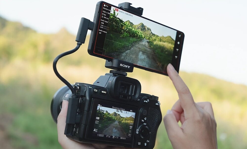 Osoba obsługuje aparat fotograficzny, do którego przymocowany jest smartfon marki SONY, wyświetlający obraz z kamery. W tle widoczny jest krajobraz z zielenią i górami.