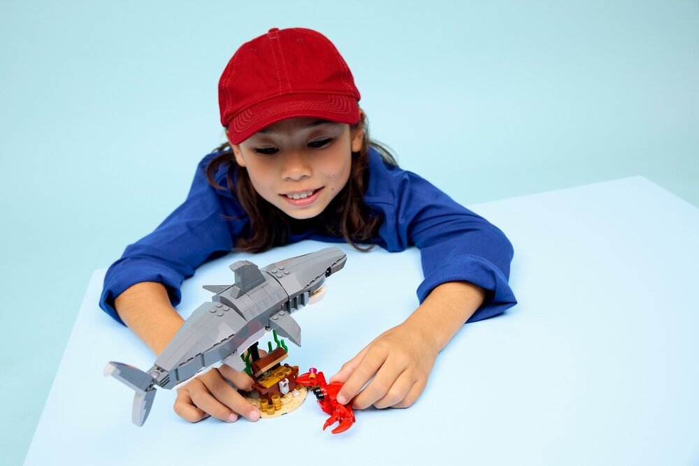 Dziecko w czerwonej czapce bawi się zestawem LEGO, który przedstawia szarego rekina i skrzynię ze skarbem. Na stole znajduje się również mały czerwony krab.