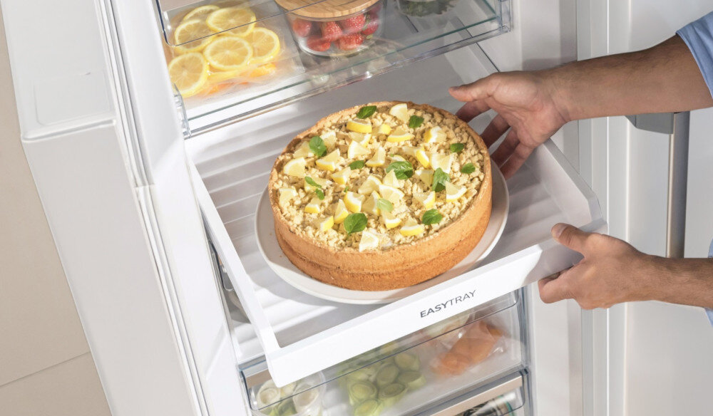 Osoba wkłada ciasto na półkę oznaczoną napisem \'EASYTRAY\' w zamrażarce. W tle widoczne są inne produkty spożywcze, takie jak cytryny i winogrona.