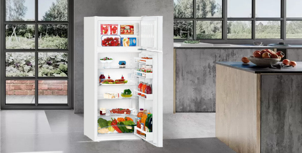Biała lodówka z otwartymi drzwiami, wypełniona różnorodnymi produktami spożywczymi, stoi w nowoczesnej kuchni. W tle widoczne są duże okna i szare ściany.