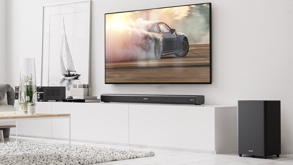 W nowoczesnym salonie znajduje się duży telewizor z wyścigowym samochodem na ekranie, a pod nim soundbar marki SHARP. Obok stoi bezprzewodowy subwoofer, a na białej szafce widoczne są różne urządzenia elektroniczne.