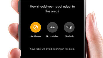 Na ekranie telefonu widoczne są trzy ikony z napisami: \'Avoid area\', \'No brush bar\', \'No climb\'. Powyżej znajduje się pytanie: \'How should your robot adapt in this area?\', a poniżej tekst: \'Your robot will avoid cleaning in this area.\'.