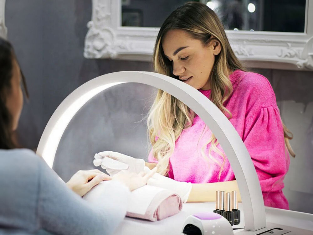 Na obrazie widać kobietę w różowej bluzce, która wykonuje manicure przy użyciu lampy LED. Na stole znajdują się buteleczki z lakierem do paznokci i urządzenie do manicure.