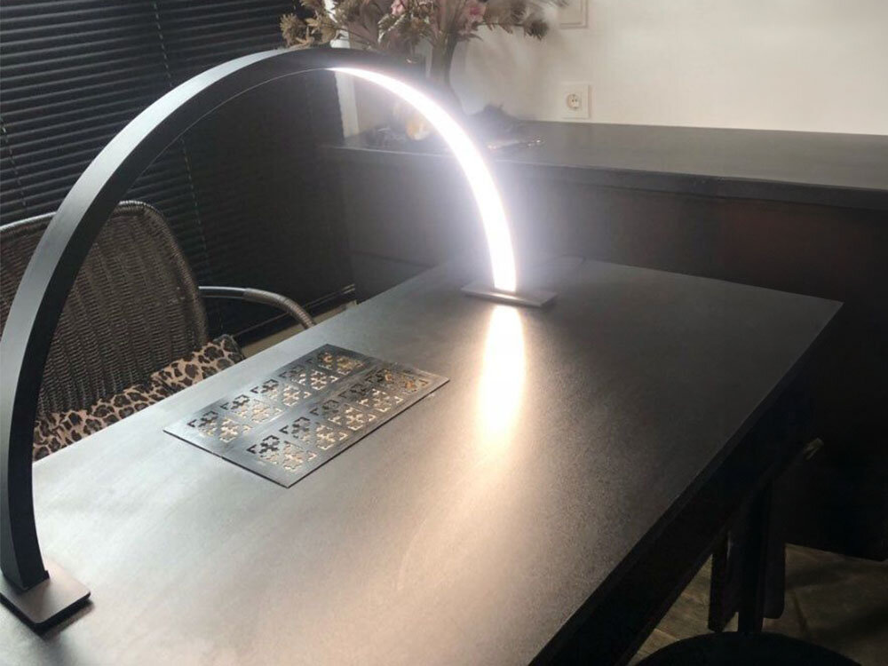 Na ciemnym stole znajduje się czarna lampa LED w kształcie łuku, emitująca jasne światło. W tle widoczny jest fotel oraz dekoracje na blacie.