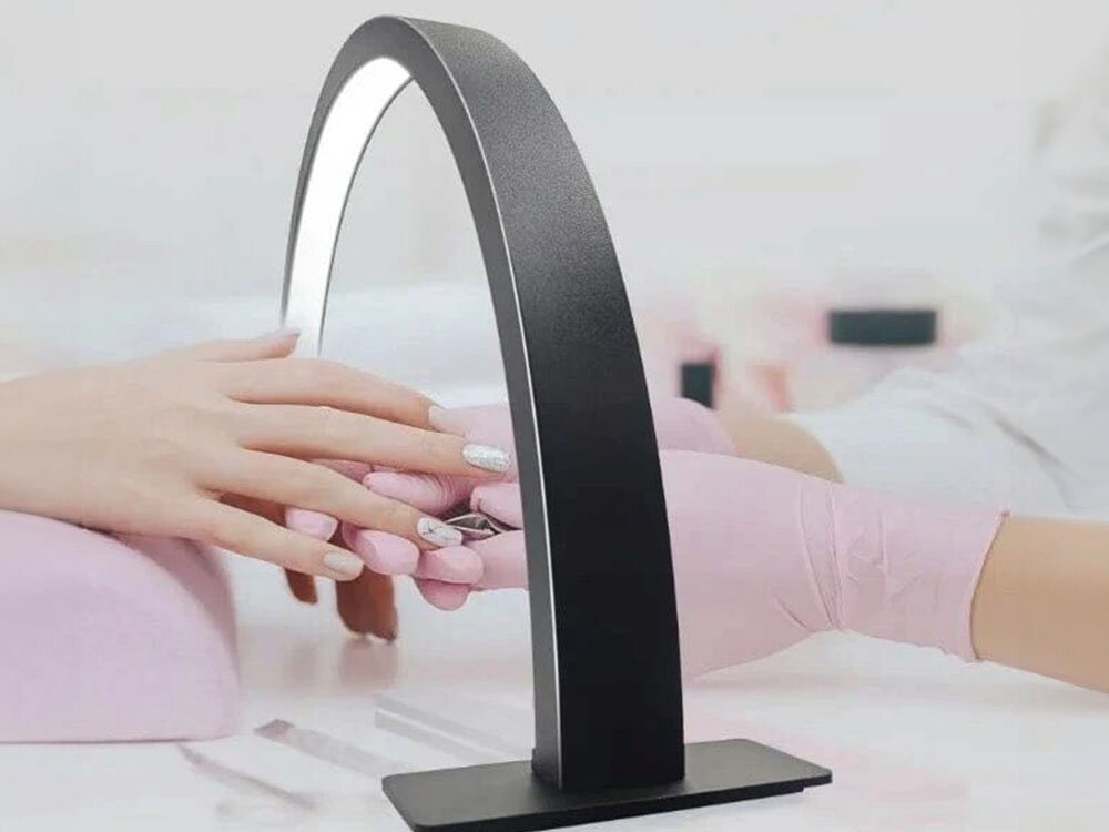 Czarna lampa LED do paznokci w kształcie łuku oświetla dłonie z manicurem. Osoba w różowych rękawiczkach trzyma dłoń klientki na różowej poduszce.
