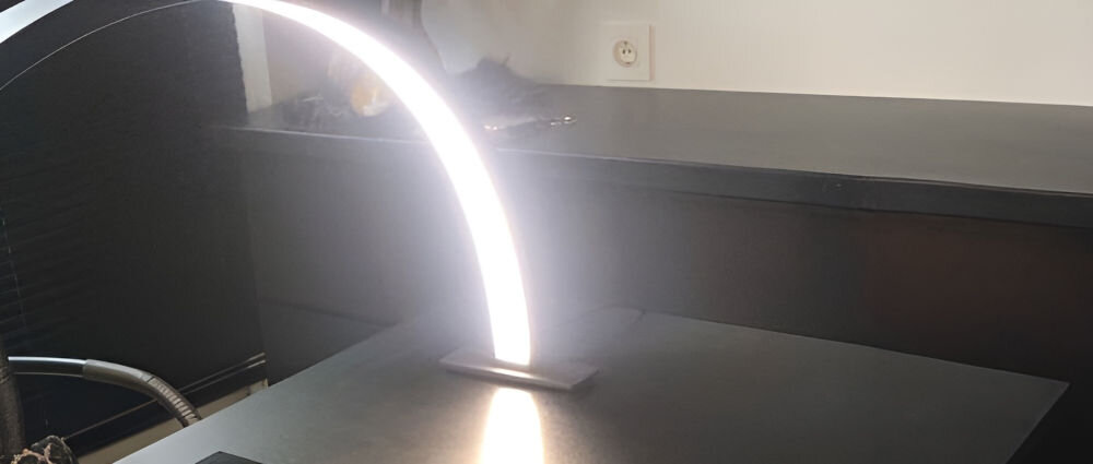 Na obrazie widoczna jest czarna lampa LED do paznokci w kształcie łuku, emitująca jasne światło. Lampa znajduje się na ciemnym blacie w pomieszczeniu z gniazdkiem elektrycznym na ścianie.