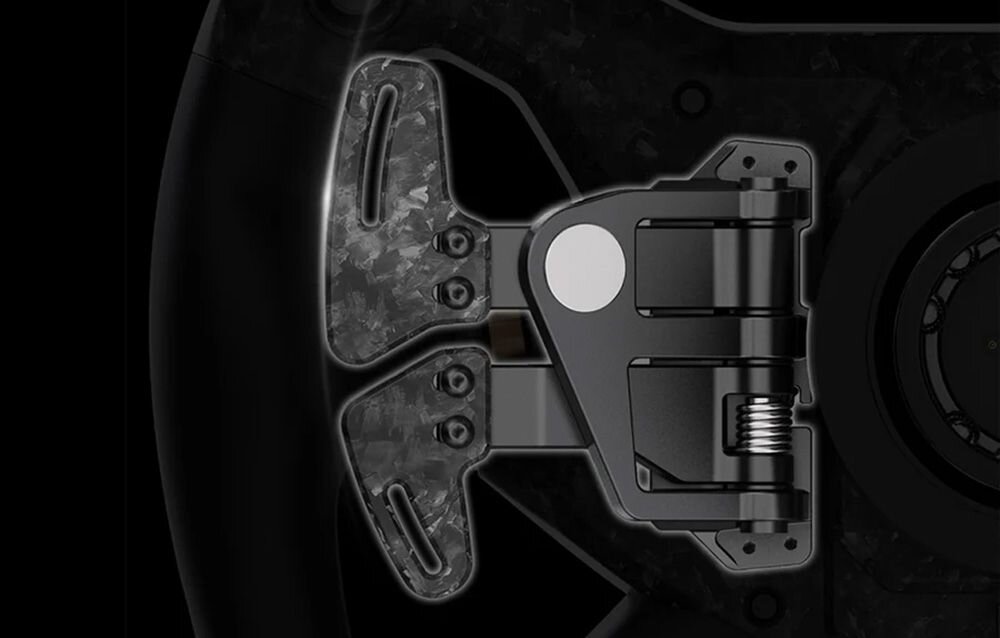Obraz przedstawia zbliżenie na mechanizm łopatkowy kierownicy wyścigowej z elementami z włókna węglowego i metalowymi częściami. Widoczna jest sprężyna oraz okrągły, srebrny element na środku.