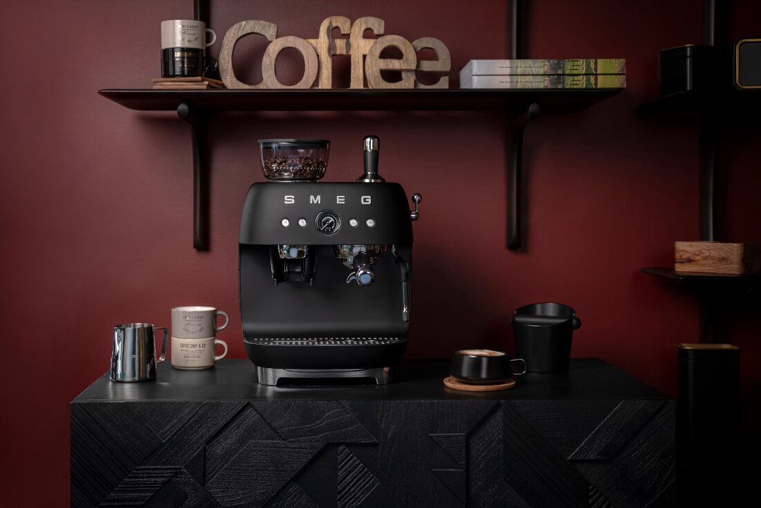 Czarny matowy ekspres do kawy z logo SMEG stoi na ciemnym blacie. W tle znajduje się półka z napisem \'Coffee\' oraz różnymi przedmiotami.