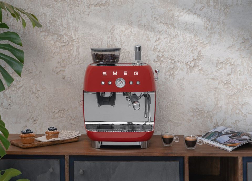 Czerwony ekspres do kawy z logo SMEG stoi na blacie obok dwóch filiżanek espresso i dwóch babeczek. W tle widoczna jest jasna, teksturowana ściana oraz roślina.