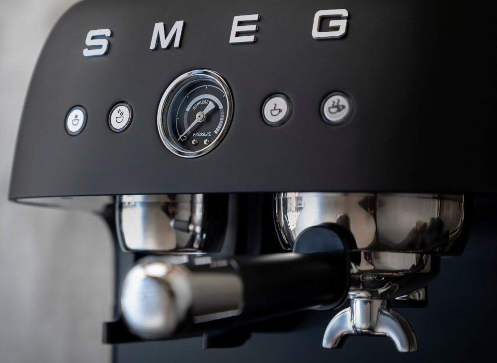 Czarny matowy ekspres do kawy z logo SMEG, posiadający manometr z napisem \'ESPRESSO PRESSURE\' oraz trzy przyciski z ikonami filiżanek.
