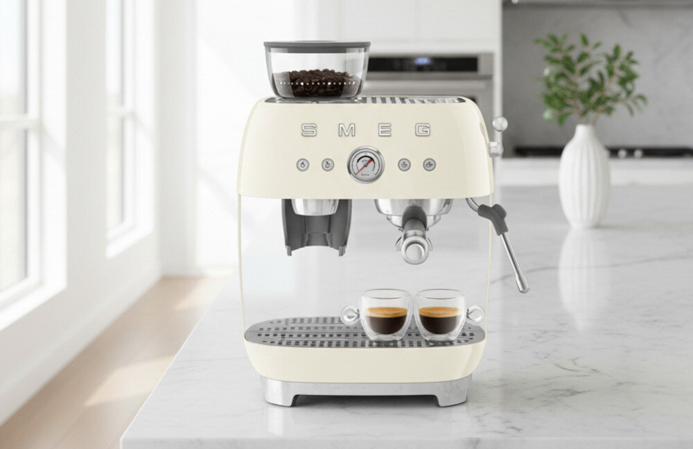 Kremowy ekspres do kawy z logo SMEG, na blacie kuchennym, z dwoma filiżankami espresso na tacy ociekowej. W tle widać jasne wnętrze kuchni z rośliną w wazonie.