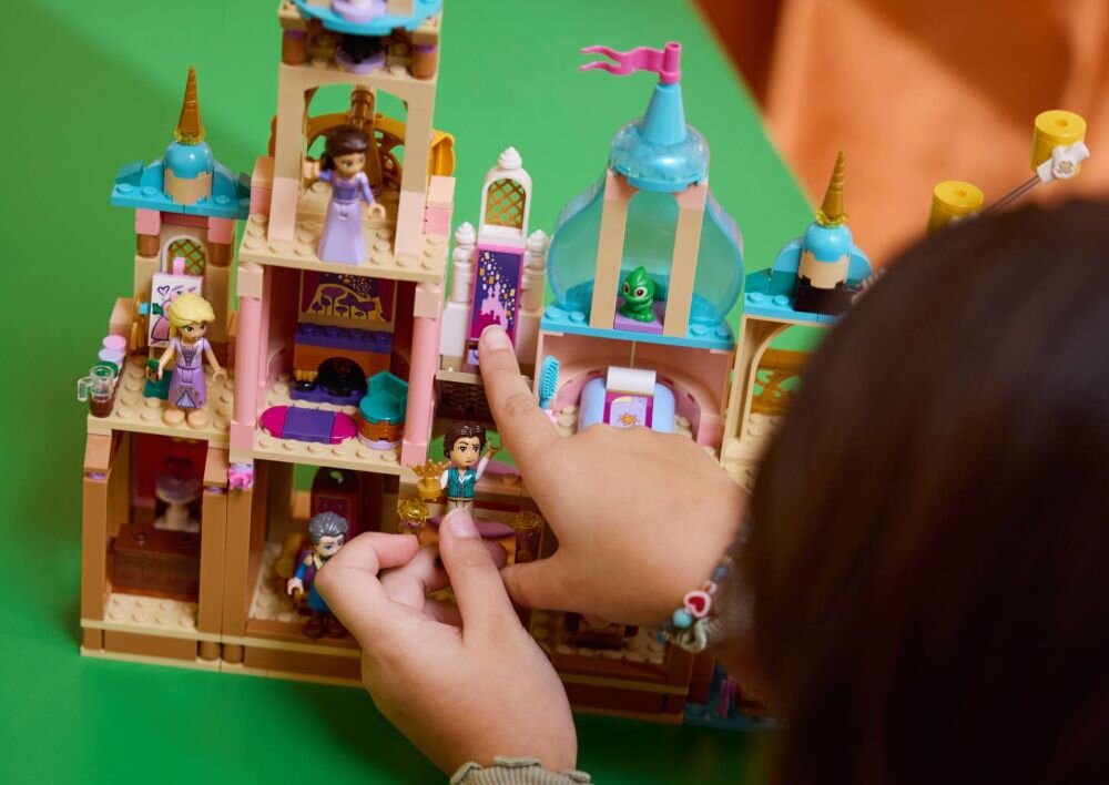 Na zielonym tle znajduje się zestaw LEGO przedstawiający zamek z figurkami postaci. Dziecko bawi się zestawem, wskazując na jedną z figurek.