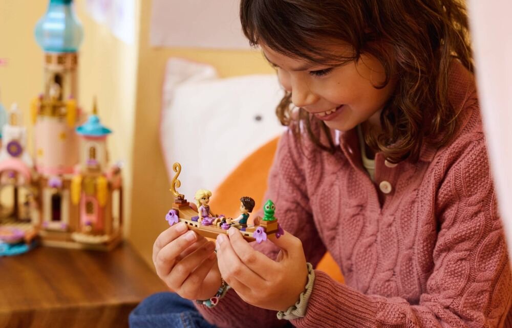 Dziecko trzyma w rękach zestaw LEGO z figurkami, a w tle widoczny jest zamek z klocków LEGO. Zamek ma różowe i niebieskie elementy oraz wieżę.
