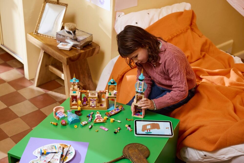 Dziecko bawi się zestawem klocków LEGO na zielonym stoliku, obok leży tablet z wyświetlonym obrazem zamku. W tle widać pomarańczowy koc na łóżku i drewniany stolik z książkami.
