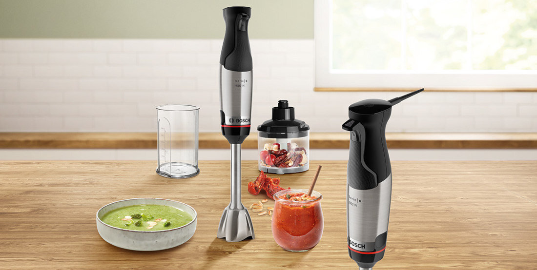 Na drewnianym blacie znajduje się blender ręczny z logo BOSCH, pojemnik z pokrywką, szklanka z czerwonym napojem oraz miska z zieloną zupą. W tle widoczna jest jasna kuchnia z oknem.