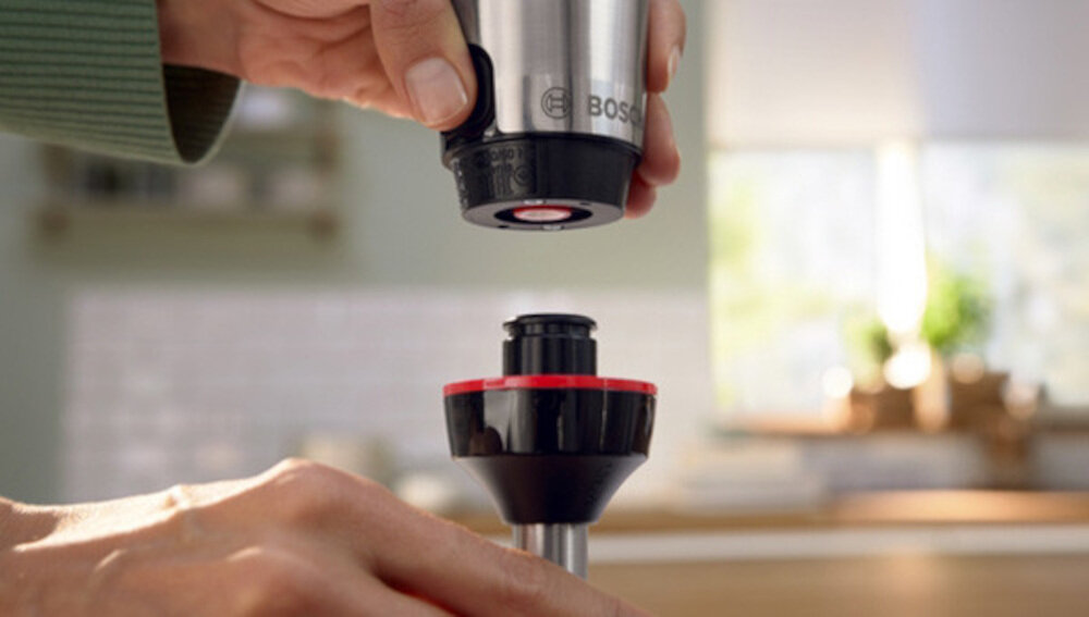 Osoba trzyma część blendera z widocznym logo BOSCH, przygotowując się do połączenia z innym elementem urządzenia. W tle znajduje się rozmyta kuchnia z jasnym oświetleniem.