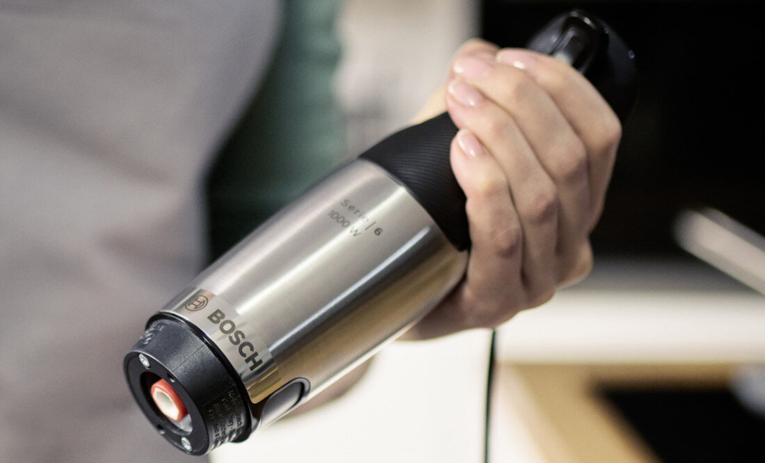 Osoba trzyma blender ręczny z metalową obudową, na której widoczne jest logo BOSCH. W tle rozmyte elementy kuchni.