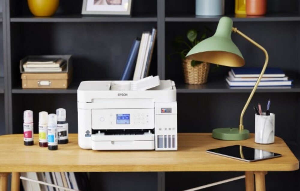 Na drewnianym stole stoi białe urządzenie wielofunkcyjne z logo EPSON, obok którego znajdują się cztery butelki z tuszem w różnych kolorach. W tle widać lampkę biurkową, tablet oraz półkę z książkami i ozdobami.