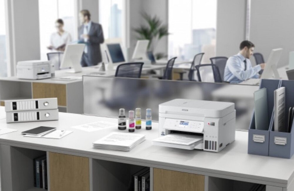 Na biurku w biurze znajduje się urządzenie wielofunkcyjne z logo EPSON, obok którego leżą butelki z tuszem i dokumenty. W tle widać osoby pracujące przy komputerach.